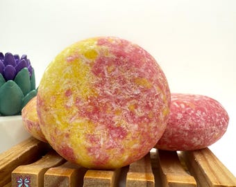May include: Two pink and yellow solid shampoo bars on a wooden rack. The bars have a speckled appearance.