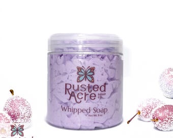 Op de afbeelding: Een doorzichtige pot gevuld met lavendel whipped soap. De pot heeft een doorzichtig deksel en een etiket met een vlinderontwerp en de woorden "Rusted Acre Soap Co." en "Whipped Soap". Decoratieve paarse bollen omringen de pot.