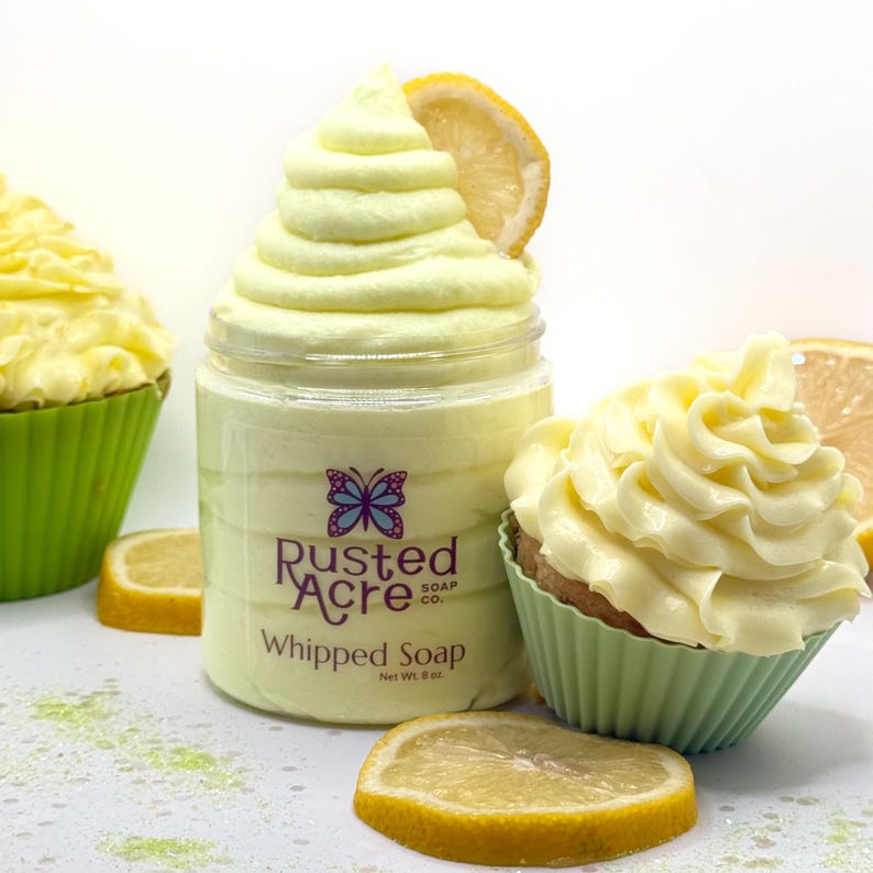 May include: A clear plastic jar of pale yellow whipped soap labeled Rusted Acre Soap Co. with a butterfly logo, topped with a swirl of soap resembling frosting and a lemon slice. Beside it sits a cupcake-shaped soap in a green liner.