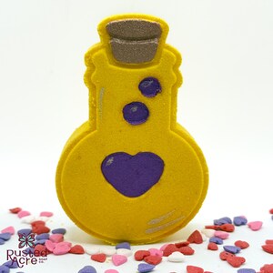 May include: A yellow bath bomb shaped like a potion bottle with a purple heart and dots. The bottle has a brown cork and is surrounded by colorful heart-shaped sprinkles. The Rusted Acre Soap Co. logo is visible.