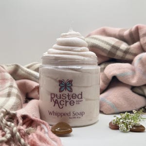 May include: A clear plastic jar filled with pale pink whipped soap, topped with a swirl. The label features a butterfly icon and the text Rusted Acre Soap Co. Whipped Soap Net Wt. 8 oz. The jar is positioned near two small brown stones.