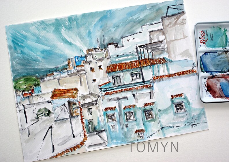 May include: Watercolor painting of a cityscape with blue and brown buildings. The buildings are clustered together and have a variety of shapes and sizes. The sky is a light blue color and there are clouds in the distance. The painting is signed "TOMYN" in the bottom right corner.