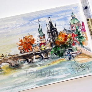 May include: Watercolor painting of a cityscape with a bridge, river, and buildings. The buildings have colorful roofs and there are trees in the foreground. The painting is signed "Tomyn" in the bottom left corner and "for love" in the bottom right corner.