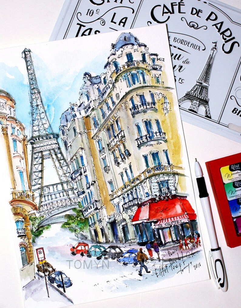 May include: Watercolor painting of a Parisian street scene with the Eiffel Tower in the background. The painting features a cafe with a red awning and a building with a yellow facade. The painting is signed "TOMYN" and dated "2018".