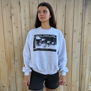 May include: A light gray sweatshirt featuring a black and white graphic of a winter scene with cabins and the text "Valley Forge Park Winter Encampment 1777-1778". The sweatshirt has long sleeves and a crew neck.