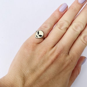 May include: A silver ring with a white alien head design. The alien has black eyes and a simple face.