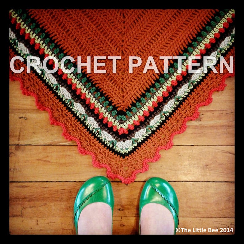 Pode incluir: Um padr&atilde;o de croch&ecirc; para um xale triangular com uma borda colorida. A borda apresenta fio vermelho, verde, branco e preto. O xale est&aacute; colocado sobre um piso de madeira.