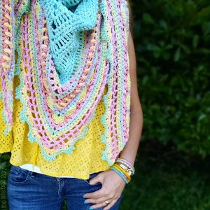 Pode incluir: Um xale de croch&ecirc; em tons de azul, rosa e amarelo. O xale tem formato triangular e um delicado padr&atilde;o de renda. O xale &eacute; usado por uma pessoa que est&aacute; vestindo uma blusa amarela e cal&ccedil;as jeans azuis.