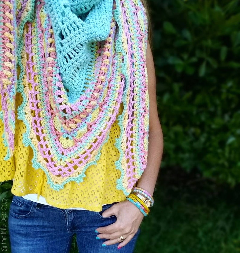 Pode incluir: Um xale de croch&ecirc; em tons de azul, rosa e amarelo. O xale tem formato triangular e um delicado padr&atilde;o de renda. O xale &eacute; usado por uma pessoa que est&aacute; vestindo uma blusa amarela e cal&ccedil;as jeans azuis.