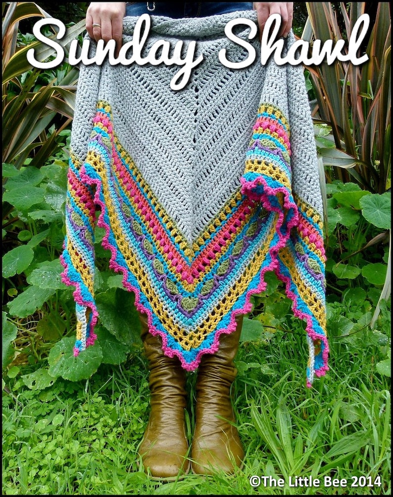 Pode incluir: Um xale de croch&ecirc; cinza com uma borda colorida em tons de rosa, amarelo, azul e verde. O xale est&aacute; drapeado sobre os ombros de uma pessoa e as pontas est&atilde;o penduradas. O xale tem uma borda festonada.