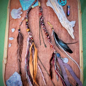 Op de afbeelding: Boheemse haaraccessoires met veren, kralen en lederen accenten, gerangschikt op een houten dienblad. Inclusief een vredesteken, kristallen en een kaart met tekst over de maan.