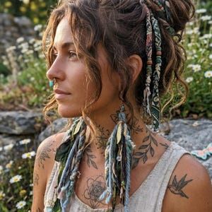 May include: A woman with curly brown hair styled in a bun, adorned with fabric and beaded hair accessories. She wears long, dangling earrings with blue and green accents. A nose ring and tattoos on her neck and shoulder are visible.