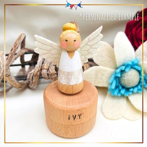 May include: A wooden angel figurine with white wings and a white dress, standing on a wooden music box with the name "IVY" engraved on it. The angel has a yellow bun and a painted face with pink cheeks.