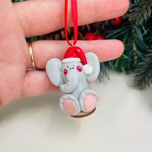 May include: A grey elephant Christmas ornament with a red Santa hat and ribbon. The elephant has pink accents on its ears and feet. The ornament is held by a hand.