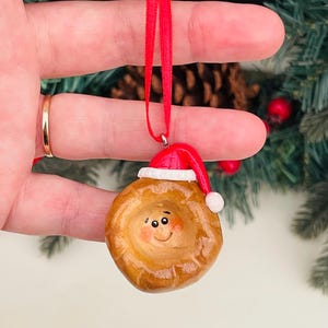 May include: A festive Christmas ornament resembling a pie, featuring a smiling face and a Santa hat. The brown ornament has a white hat trim and a red ribbon for hanging. A charming festive decoration.