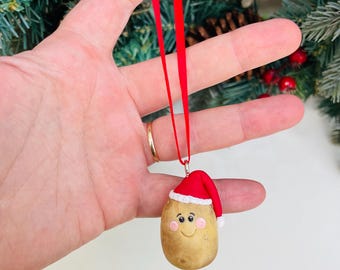 May include: A brown potato ornament with a red Santa hat and a white trim. The potato has a smiling face with pink cheeks and black eyes. The ornament is hanging from a red ribbon.