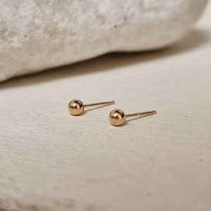 以下が含まれることがあります： 金色のボールスタッドピアスのペア。ピアスは小さくシンプルで、滑らかで丸い形をしています。