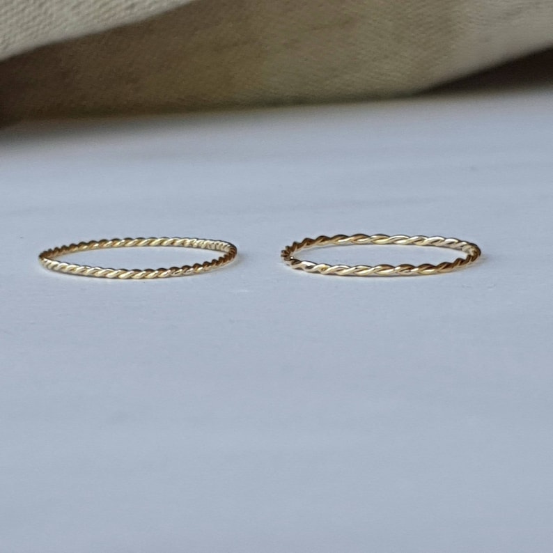 Op de afbeelding: Twee dunne gouden ringen met een gevlochten ontwerp. De ringen liggen op een wit oppervlak.
