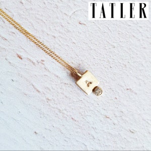 Puede incluir: Un collar de cadena de oro con un colgante cuadrado que presenta la letra "A" y un pequeño diamante. El colgante está unido a la cadena con un delicado lazo.