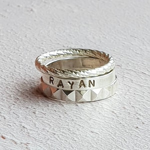 Puede incluir: Tres anillos de plata apilados uno encima del otro. El anillo superior tiene un diseño de cuerda trenzada. El anillo del medio tiene el nombre "RAYAN" grabado. El anillo inferior tiene un patrón texturizado en forma de diamante.