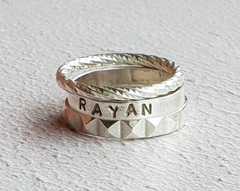 May include: Three silver rings stacked on top of each other. The top ring has a twisted rope design. The middle ring has the name "RAYAN" engraved on it. The bottom ring has a textured, diamond-shaped pattern.