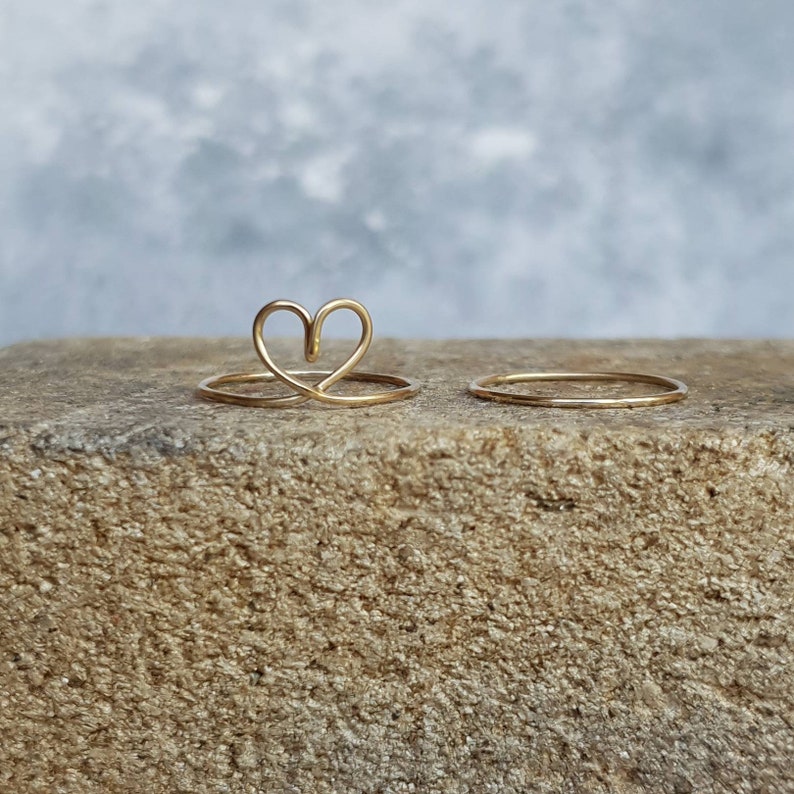 Puede incluir: Dos anillos de oro sobre una superficie rugosa y marr&oacute;n. Un anillo es una banda simple, el otro tiene forma de coraz&oacute;n.