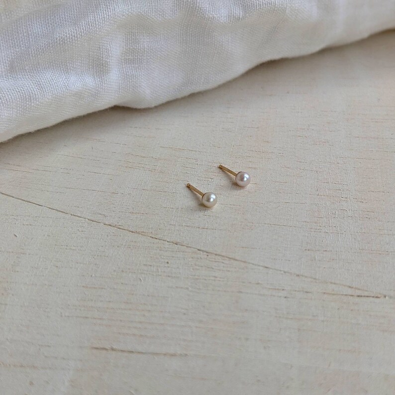 May include: Two small pearl stud earrings on a light wood surface. The earrings are gold and have a small pearl in the center.