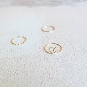 May include: Three gold rings on a white background. One ring is a simple band, one is a beaded band, and one is a heart-shaped ring.