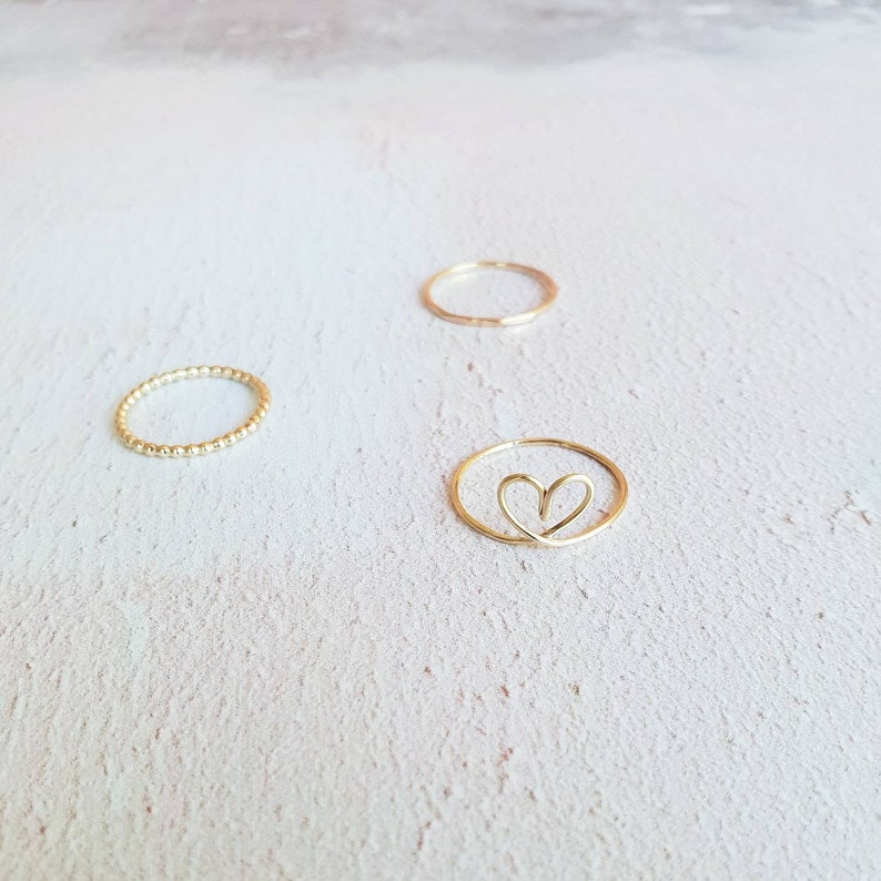May include: Three gold rings on a white background. One ring is a simple band, one is a beaded band, and one is a heart-shaped ring.