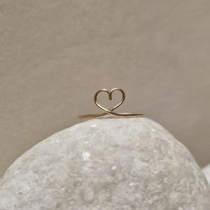 May include: A delicate gold ring with a simple heart design. The ring is sitting on a white rock.