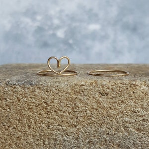 May include: Two gold rings on a rough, brown surface. One ring is a simple band, the other is shaped like a heart.