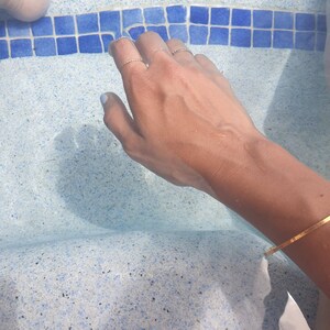 Op de afbeelding: Een hand met gouden ringen aan de vingers is ondergedompeld in een blauw-wit betegeld zwembad. De hand draagt een gouden armband.