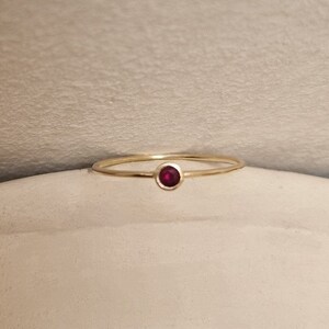 May include: A delicate gold ring featuring a small, round, deep red gemstone. The ring has a thin band and a simple, elegant design. The gemstone is set in a bezel setting, and the ring is displayed on a neutral surface.