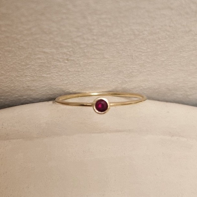 May include: A delicate gold ring featuring a small, round, deep red gemstone. The ring has a thin band and a simple, elegant design. The gemstone is set in a bezel setting, and the ring is displayed on a neutral surface.