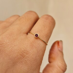 May include: A delicate gold ring featuring a small, round, deep red gemstone. The ring has a thin band and a simple, minimalist design. The gemstone is set in a bezel setting, adding a touch of elegance to the piece.