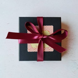 K&ouml;nnte beinhalten: Eine schwarze Geschenkbox mit einem burgunderfarbenen Band, das zu einer Schleife gebunden ist. Die Box hat einen Aufkleber mit einem Chevron-Muster und dem Wort "Hand" darauf.