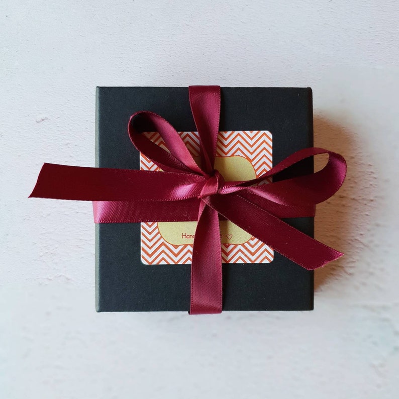 K&ouml;nnte beinhalten: Eine schwarze Geschenkbox mit einem burgunderfarbenen Band, das zu einer Schleife gebunden ist. Die Box hat einen Aufkleber mit einem Chevron-Muster und dem Wort "Hand" darauf.