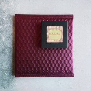 K&ouml;nnte beinhalten: Ein burgunderfarbener gepolsterter Umschlag mit einer kleinen schwarzen Box darauf. Die Box hat einen goldenen Aufkleber mit dem Text "TheChokerStory" und "Handmade with &hearts;".