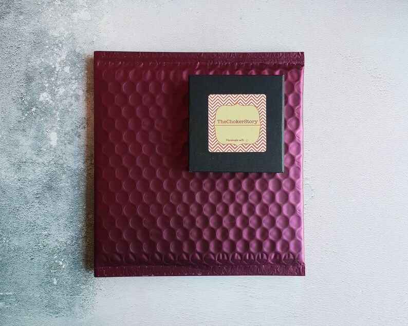 K&ouml;nnte beinhalten: Ein burgunderfarbener gepolsterter Umschlag mit einer kleinen schwarzen Box darauf. Die Box hat einen goldenen Aufkleber mit dem Text "TheChokerStory" und "Handmade with &hearts;".