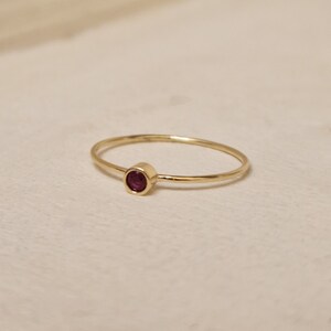 May include: A delicate gold ring featuring a small, round, deep red gemstone. The ring has a thin band and a simple, elegant design. The gemstone is set in a bezel setting, adding a touch of color and sophistication.
