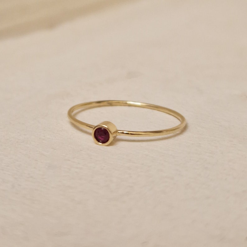 May include: A delicate gold ring featuring a small, round, deep red gemstone. The ring has a thin band and a simple, elegant design. The gemstone is set in a bezel setting, adding a touch of color and sophistication.