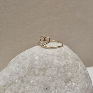 May include: A delicate gold ring with a simple heart design. The ring is shown on a white rock.
