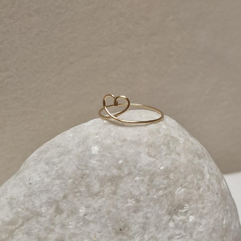 May include: A delicate gold ring with a simple heart design. The ring is shown on a white rock.
