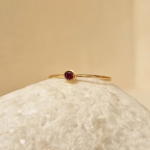 May include: A slender gold ring with a small, round ruby gemstone. The ring has a thin, polished band, and the ruby is set in a simple bezel. The background is a soft, neutral colour, which accentuates the ring's elegant design.