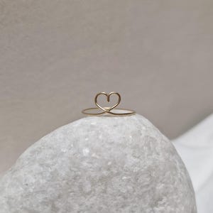 May include: A delicate gold ring with a simple heart design. The ring is sitting on a white stone.
