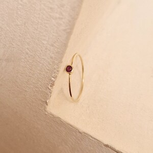 May include: A delicate gold ring featuring a small, round, deep red gemstone. The ring has a thin band and a minimalist design, showcasing the gemstone prominently. The gold band contrasts with the rich color of the stone.
