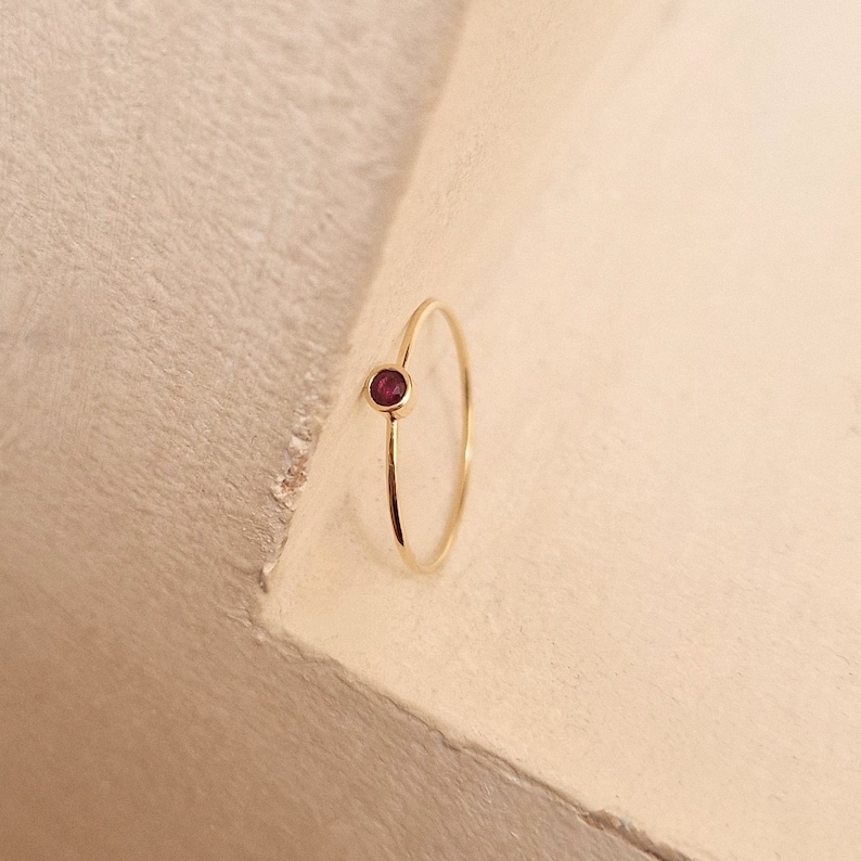 May include: A delicate gold ring featuring a small, round, deep red gemstone. The ring has a thin band and a minimalist design, showcasing the gemstone prominently. The gold band contrasts with the rich color of the stone.