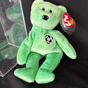 May include: A green plush bear with a soccer ball emblem on its chest. The bear has black eyes and a black nose. A red and white tag is attached to the ear. The bear is a collectible item.