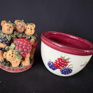 Puede incluir: Una figura decorativa de tres osos marrones sosteniendo racimos de uvas y frambuesas. Junto a ella, un cuenco de cerámica blanco con interior rojo y bayas pintadas.