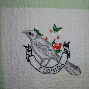 Puede incluir: Bloque de colcha blanco bordado con un pájaro gris, flores rojas y hojas verdes. El pájaro está posado en una cinta negra con la palabra "FLORIDA" bordada en ella.